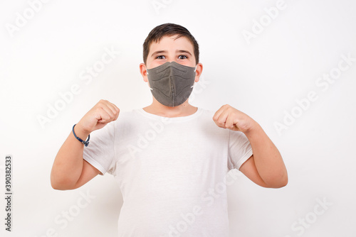 Portrait of Caucasian young boy wearing medical mask standing against white background looks with excitement at camera, keeps hands raised over head, notices something reacts on sudden news.