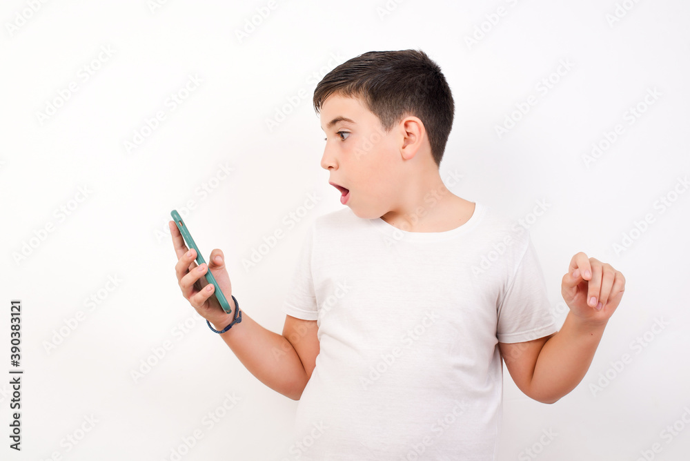 Happy cheerful Caucasian young boy standing against white background receiving good news via e-mail and celebrating success while standing and looking at mobile phone. Happy cheerful Caucasian young boy standing against white background receiving good news via e-mail and celebrating success while standing and looking at mobile phone.