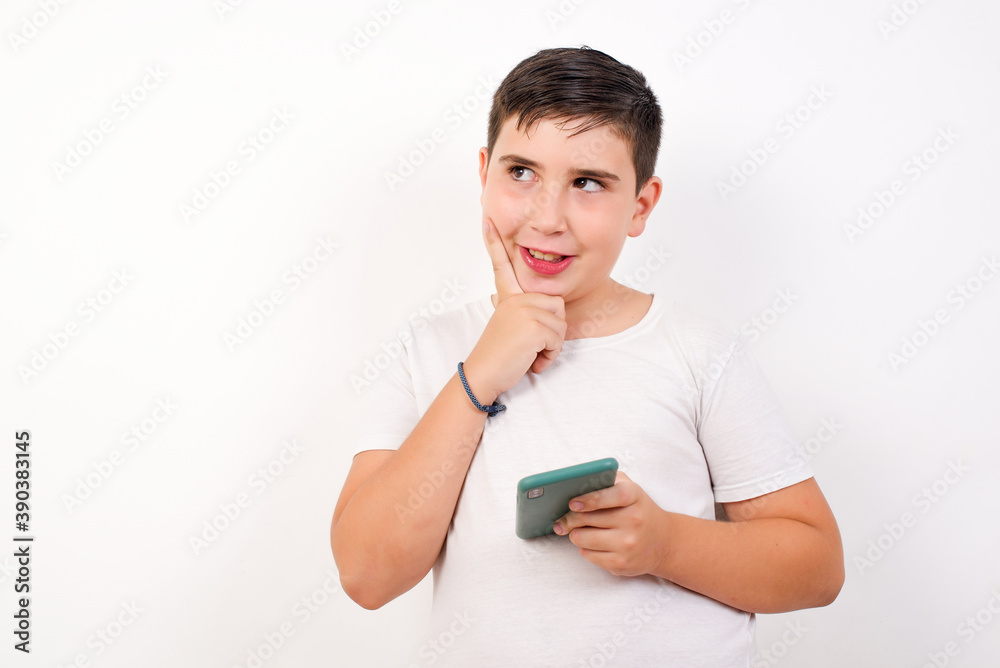 Image of a thinking dreaming Caucasian young boy standing against white background using mobile phone and holding hand on face. Taking decisions and social media concept. Image of a thinking dreaming Caucasian young boy standing against white background using mobile phone and holding hand on face. Taking decisions and social media concept.