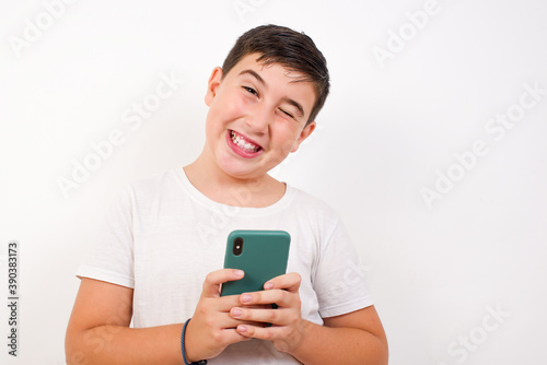 Pleased Caucasian young boy standing against white background using self phone and looking and winking at the camera. Flirt and coquettish concept. © Jihan