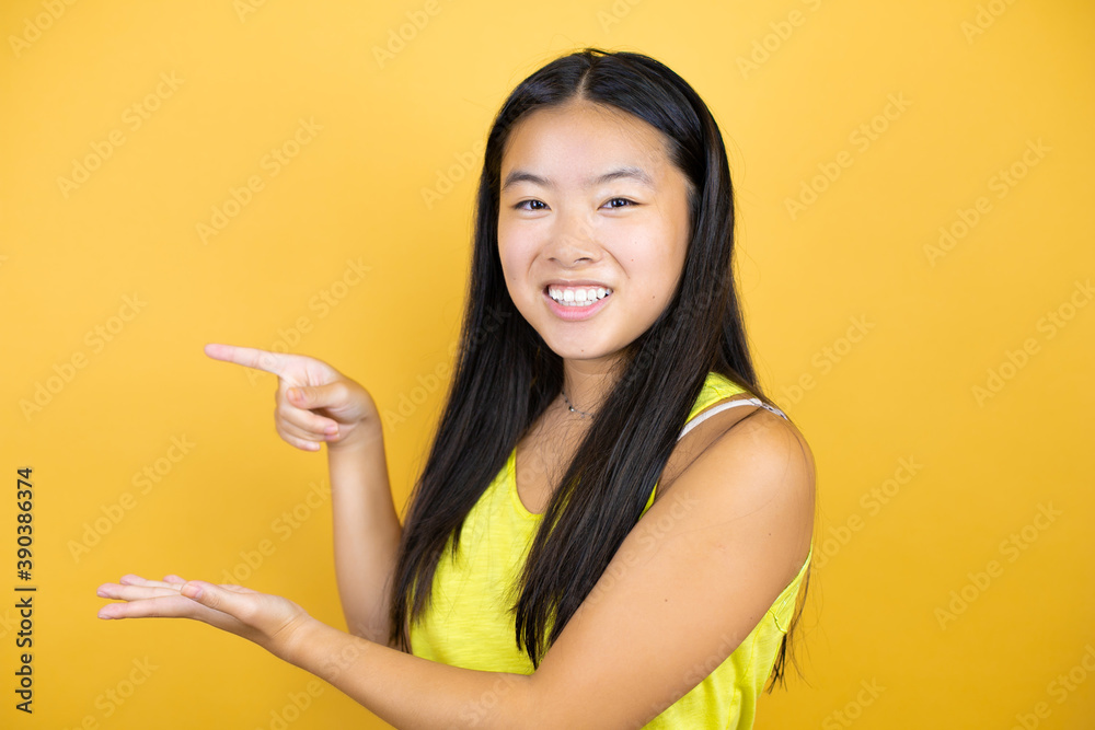 Young beautiful asian woman over yellow isolated background surprised, showing and pointing something that is on her hand Young beautiful asian woman over yellow isolated background surprised, showing and pointing something that is on her hand