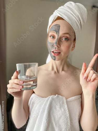 close-up of a woman with a towel on her head and a clay mask on her face. She points her finger up and holds a glass of clear water.