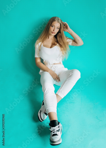 Luxury blonde in white sportswear posing in the studio on a turquoise background.