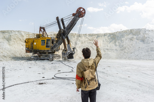the traveler stands in front of a large excavator. a young man is in the middle of an industrial quarry with white sand.