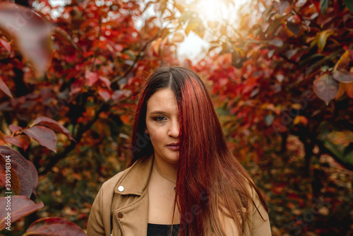 Close up young woman portrait looking at the camera with red hair covering half face in a red field with fallen leaves and sunligtht. Autumn season, color, nature concept.