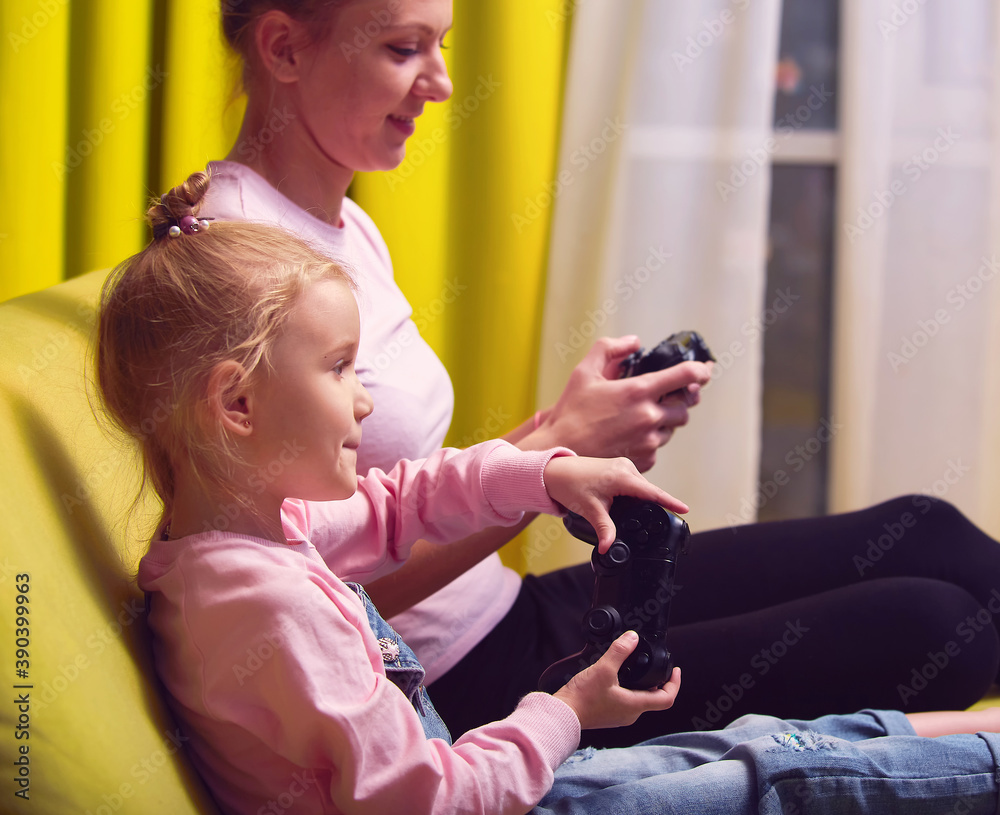 Fototapeta premium Happy family together. Mother and her child girl playing video games sit on couch ahd hold gamepad. People having fun at home