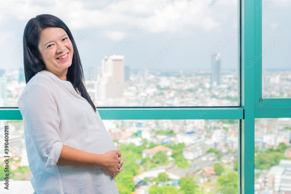 Fototapeta premium Asian pregnant woman touching her belly with hands and thinking of future. She taking a break pensive standing near condo window.
