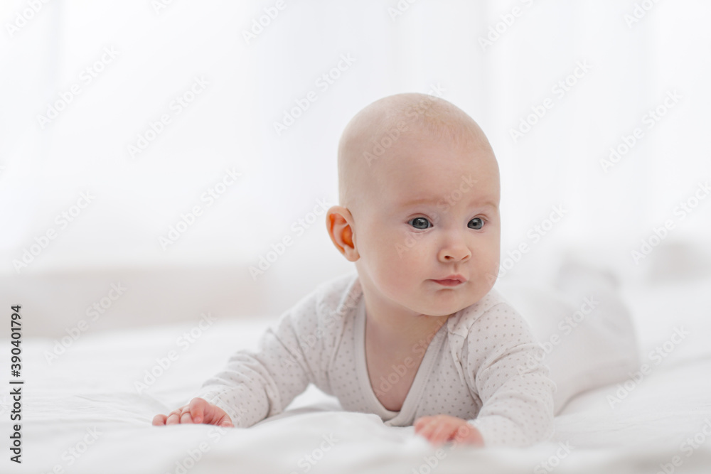 Portrait of a cute beautiful baby with gray eyes in white pajamas on the bed against the window.