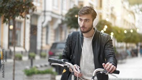 Slow Motion Young man stands on the street with bicycle and listen to music. Bike courier with backpack and smartphone, he is in a good mood, he smiles and enjoys life photo