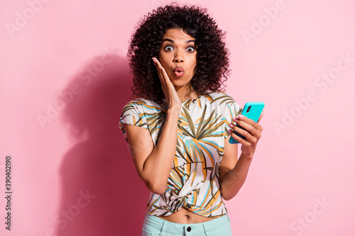 Photo portrait of surprised woman holding phone in one hand touching face cheek isolated on pastel pink colored background
