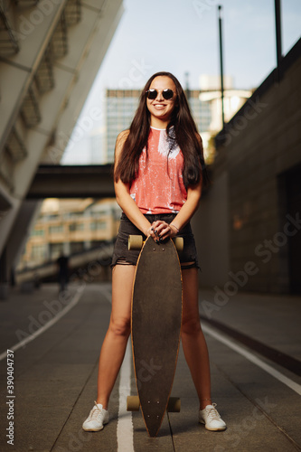 Beautiful sexy young girl in short shorts posing  with skateboard in city on sunset background