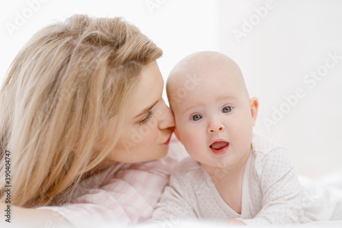 Young beautiful Caucasian blonde mom lies on the bed with her baby daughter. White light home interior.