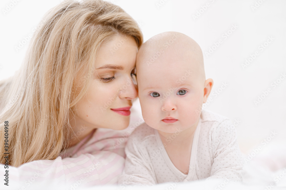 Young beautiful Caucasian blonde mom lies on the bed with her baby daughter. White light home interior. Young beautiful Caucasian blonde mom lies on the bed with her baby daughter. White light home interior.