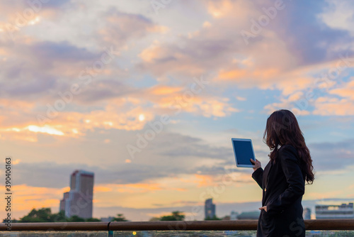 Asian businesswoman holding digital tablet, reading, touching the screen and sunset sky background.