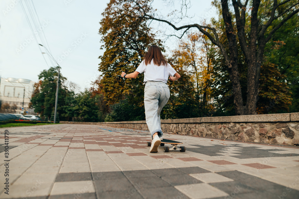 Fototapeta premium View from the back, a woman rides a longboard, pushes off the ground, photo in motion. Woman skater walks on a longboard around the city