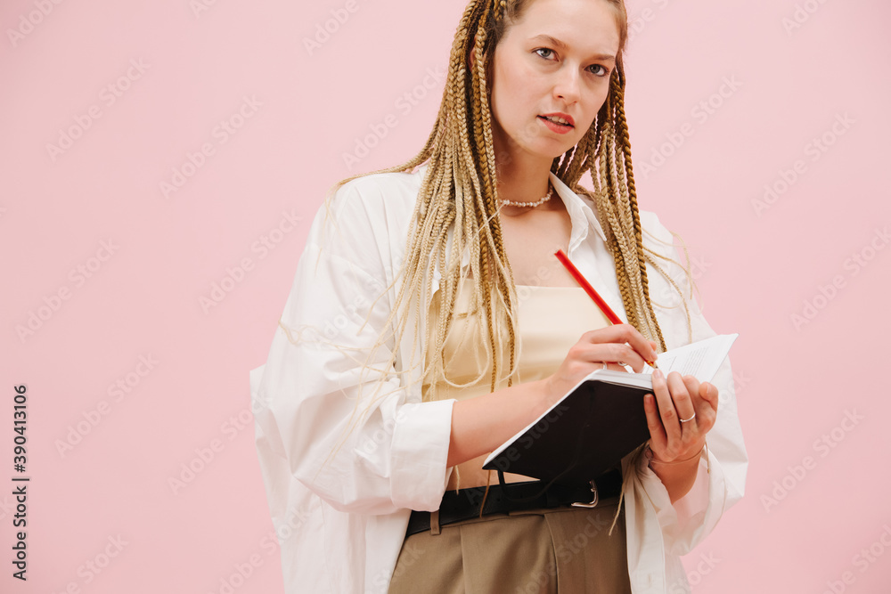 Keen young woman with stylish blond afro braids writing in a notebook over pink Keen young woman with stylish blond afro braids writing in a notebook over pink