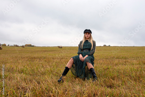A girl with blonde hair , wearing a black cap, green chiffon dress and rough boots sits in a mown field. © FO_DE