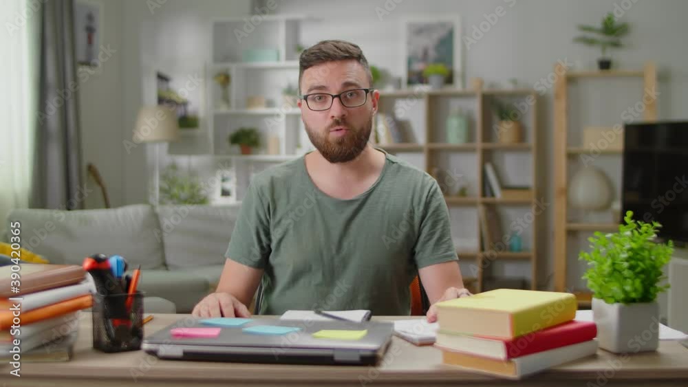 A young man sitting at a desk at home talks to the camera. The man is vlogging at home in the living room. A young man sitting at a desk at home talks to the camera. The man is vlogging at home in the living room.