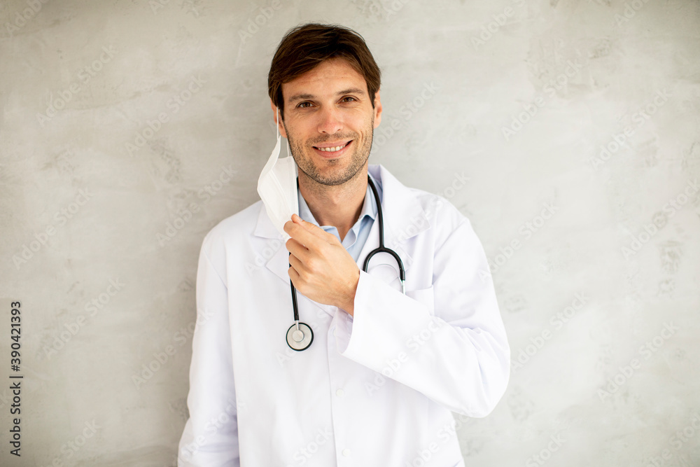 Attractive young male doctor taking off his surgical mask while standing by the wall Attractive young male doctor taking off his surgical mask while standing by the wall