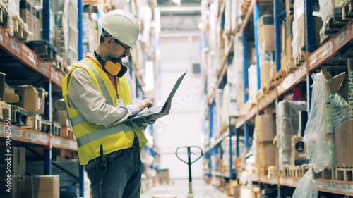 Storeman in uniform typing on laptop among warehouse shelves