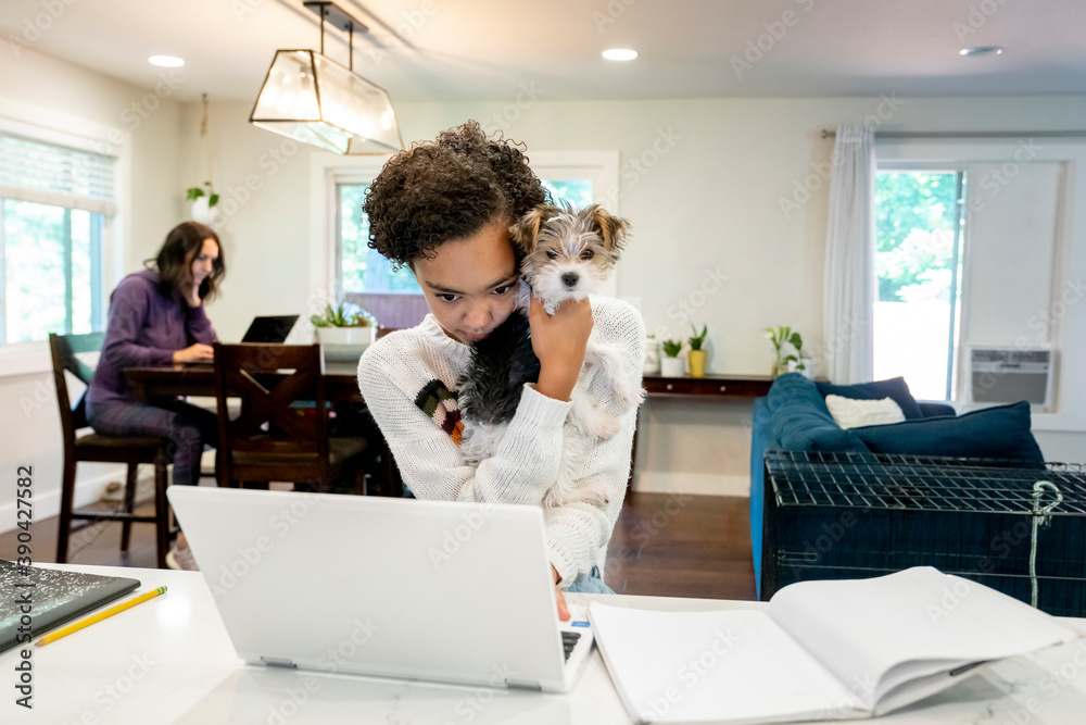 Teen girl doing remote school from home kitchen with laptop and wireless headphones while holding cute puppy, mom working in background