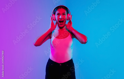 Listening to music with headphones. Young caucasian woman's portrait on gradient blue-purple studio background in neon. Concept of youth, human emotions, facial expression, sales, ad. Beautiful model.
