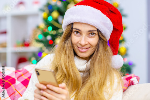 A young woman in a Santa hat sits on the couch at home with a teleon in her hands against the background of a Christmas tree. Online merry christmas greetings at lockdown