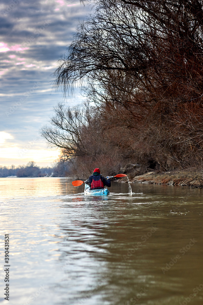 Man kayaking on blue kayak in winter river near trees. Back view