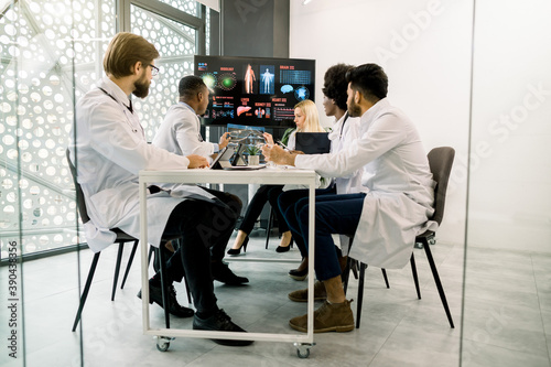 Team of qualified multiracial doctors having a medical discussion in a meeting room with big digital screen, sitting at the table and interacting each other, working with patient's CT