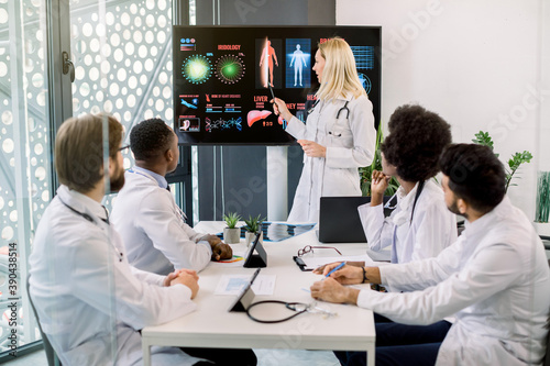 Medical education  health care and medicine concept. Team of multiethnic doctors having meeting in clinic  looking at the big digital screen  while female blond doctor sharing important information