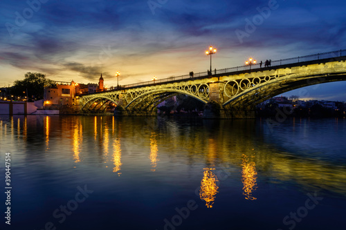 Puente de Triana sobre el río Guadalquivir en Sevilla a la puesta de sol nocturna con reflejos en el agua.