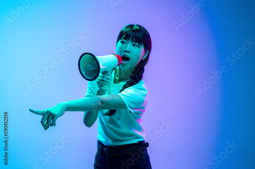 Shouting with loudspeaker. Young asian woman's portrait on gradient blue-purple studio background in neon. Concept of youth, human emotions, facial expression, sales, ad. Beautiful brunette model.