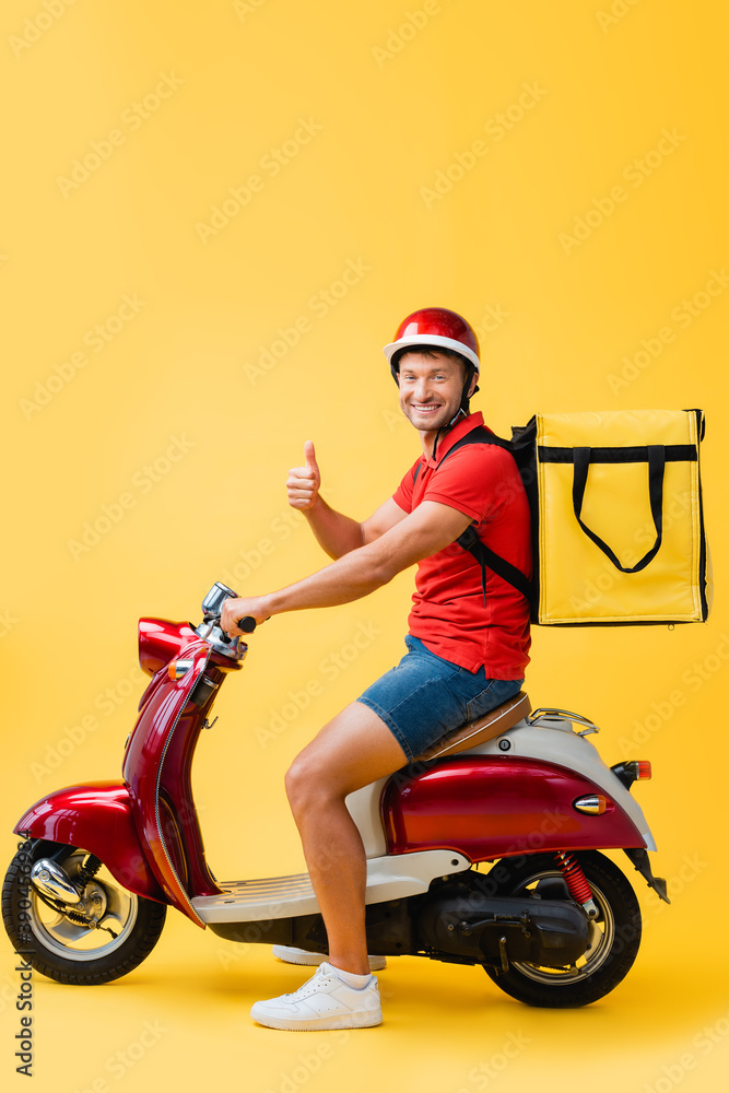 happy delivery man in helmet and backpack riding red scooter while showing thumb up on yellow happy delivery man in helmet and backpack riding red scooter while showing thumb up on yellow