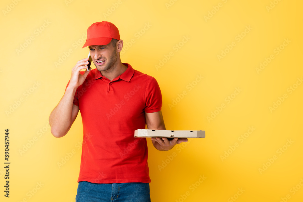 displeased delivery man in red cap holding box with tasty pizza and talking on smartphone on yellow