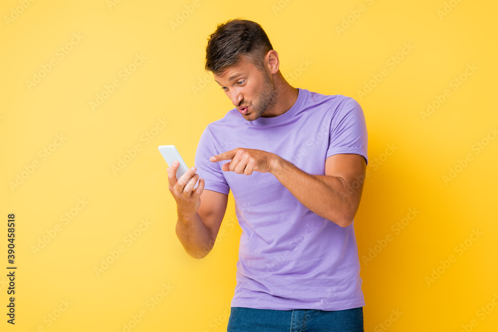 displeased man pouting lips while pointing with finger at smartphone on yellow displeased man pouting lips while pointing with finger at smartphone on yellow