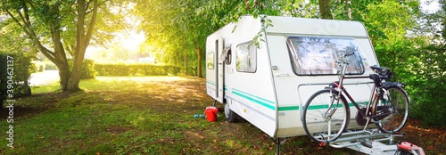 Caravan trailer with a bicycle parked on a green lawn under the trees in a camping site. France. Vacations, leisure activity, tourism, road trip, travel destinations, lifestyle. Panoramic view