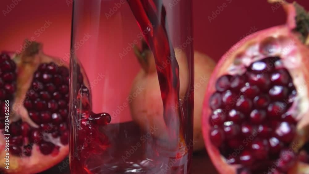 Delicious red pomegranate juice wave is flowing into a tall glass next to the halves of pomegranate on the red background in slow motion