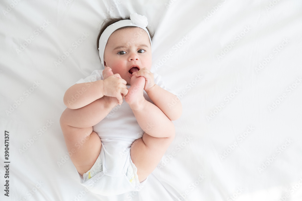 funny 6 months baby girl playing with legs, while lying in bed at home on white bedding. top view. funny 6 months baby girl playing with legs, while lying in bed at home on white bedding. top view.