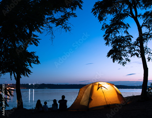 Illuminated tent and silhouettes of family sitting on the beach in the evening
