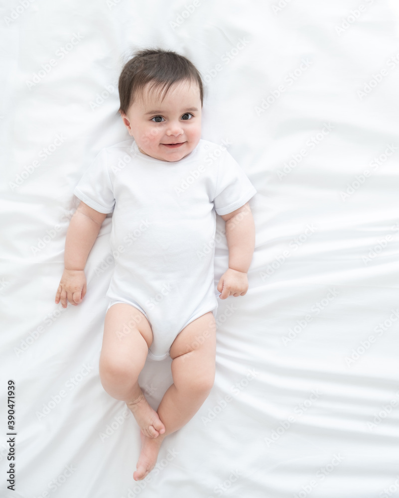 funny healthy baby girl 6 months in white bodysuit lying on bed on white bedding. top view. funny healthy baby girl 6 months in white bodysuit lying on bed on white bedding. top view.