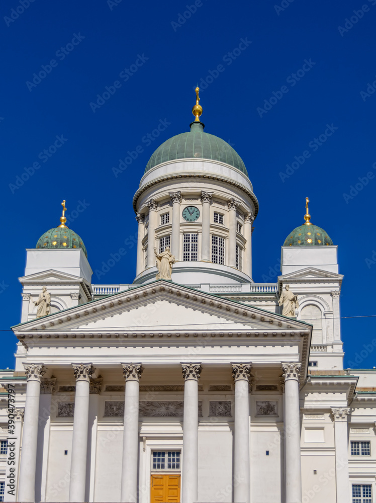 Main entrance to white Helsinki Cathedral with neoclassical green dome with a clock, surrounded by smaller domes against bright blue sky.
