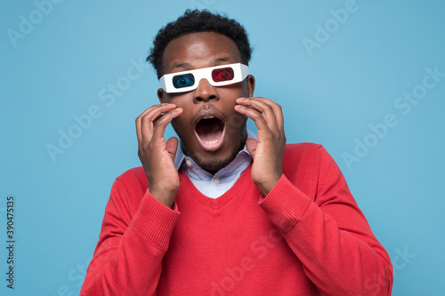 African american man in 3D glassesbeing surprised watching interesting movie on screen. Studio shot on blue wall.