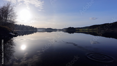 Wide panorama of lake with half solar halo in the blue sky and the reflection as a mirror image in the water. The continue ripples on the still water surface. Beautiful Scandinavia scenery.