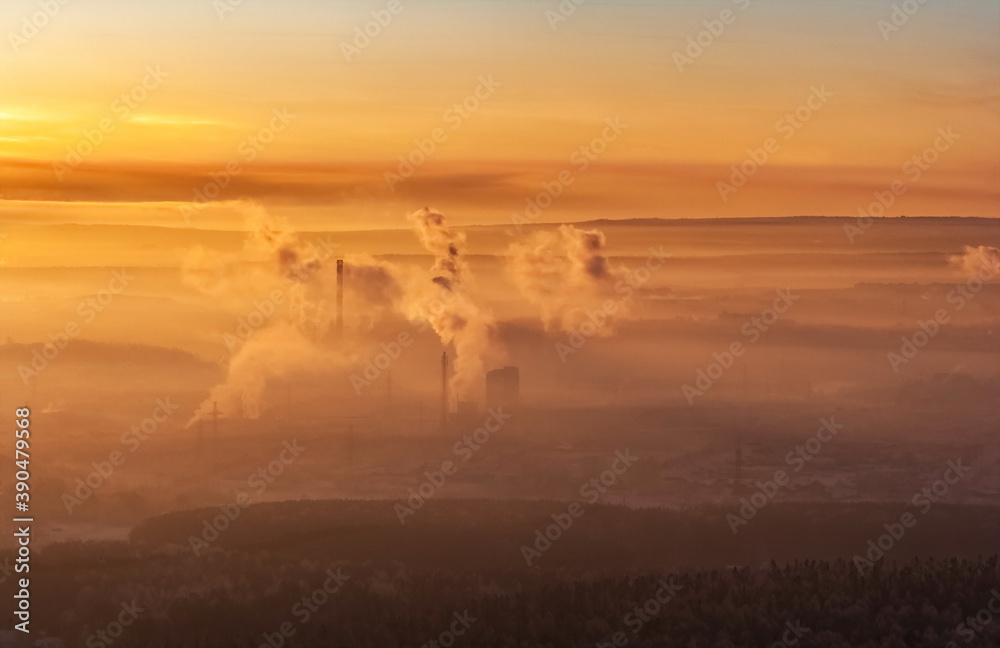 Winter landscape with forest, mountains, pipes with smoke, sky with clouds, frosty haze in the rays of the setting sun