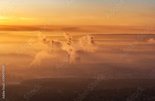 Winter landscape with forest, mountains, pipes with smoke, sky with clouds, frosty haze in the rays of the setting sun