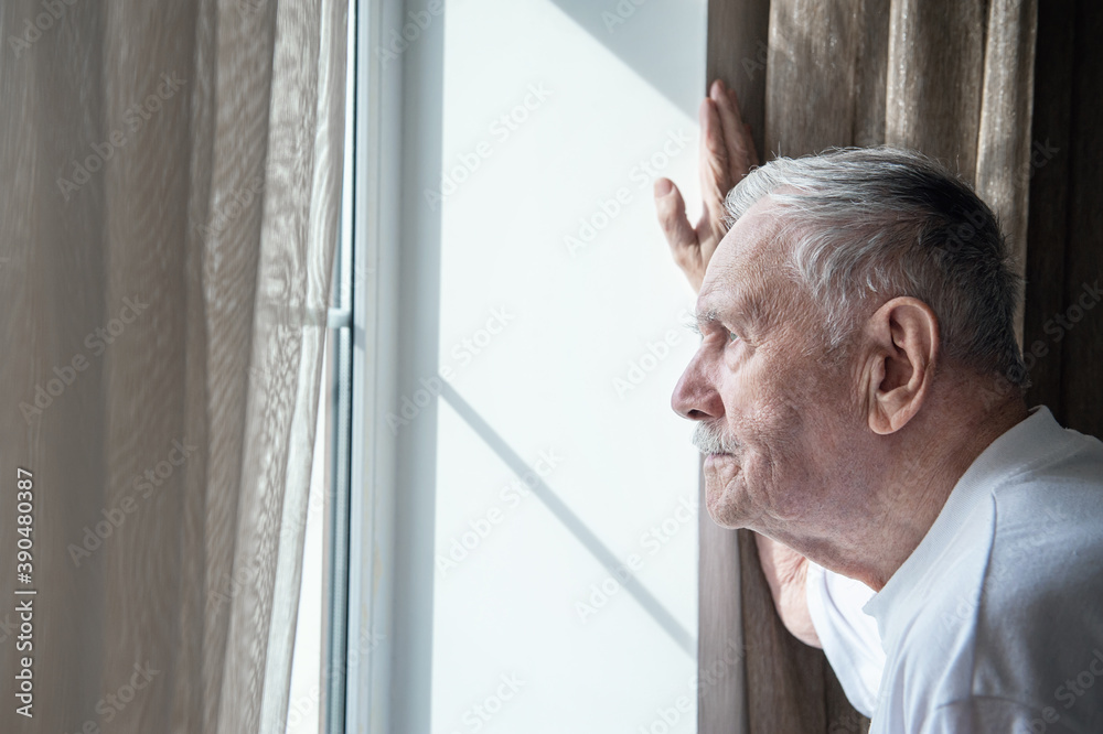 gray-haired old man, forced to remain in quarantine due to the covid-19 coronavirus, looks out the window with a disconsolate expression. Loneliness of old people. Stay home in the pandemic