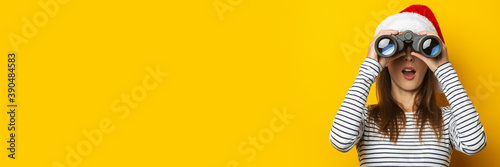 Young woman with a surprised face in a Santa Claus hat looks through binoculars on a yellow background. Banner