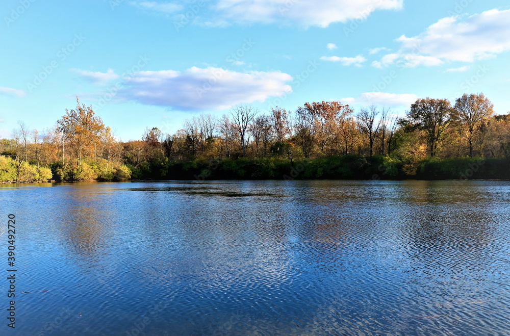 Obraz premium Beautiful autumn landscape with a view of the opposite bank of the river, clouds and trees are reflected in the blue water