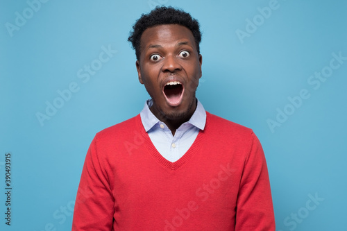 Surprised young African American man screaming in shock, keeping mouth wide open. Studio shot on blue wall.