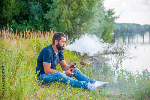 Stylish bearded vaper smoking an e-cigarette on the forest ground. Modern ways of quitting tobacco.